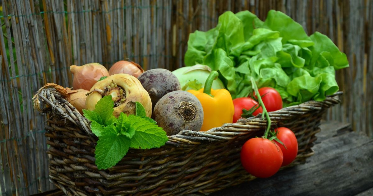 A wicker basket filled with various fresh vegetables including onions, beets, yellow bell pepper, tomatoes, lettuce, and a sprig of mint, placed against a rustic wooden background.