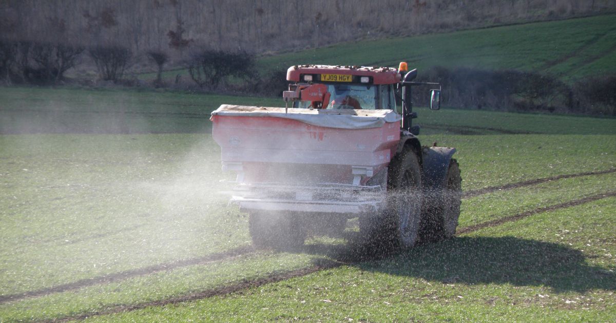 A tractor spreading fertilizer across a green field with visible spray dispersing evenly over the ground.