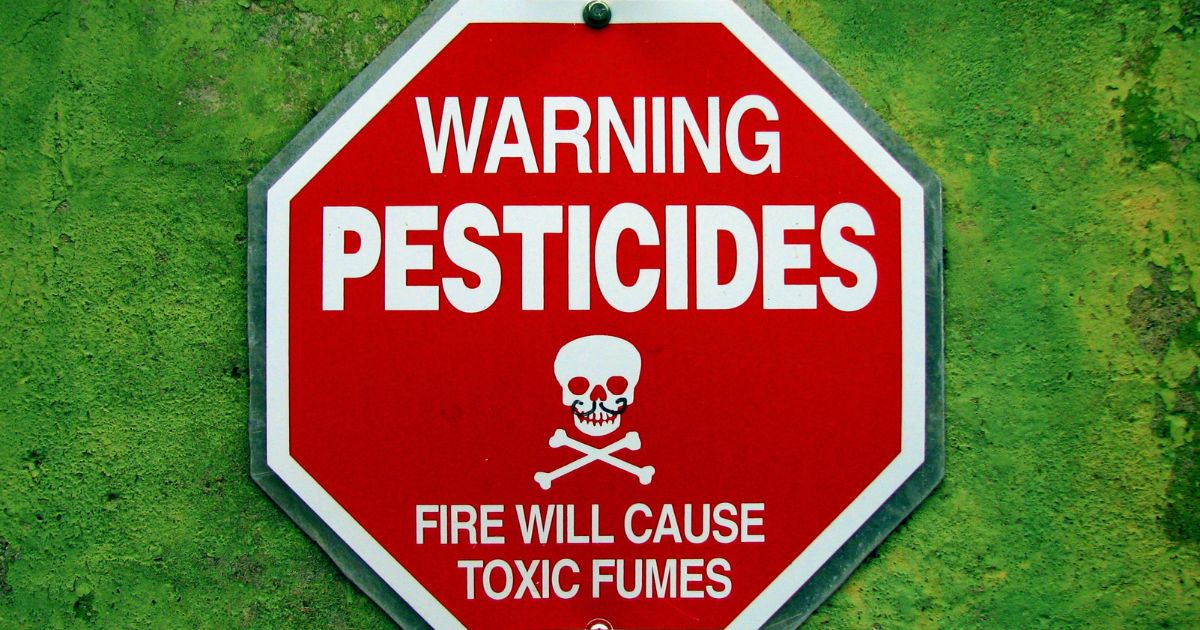 Warning sign with text 'WARNING PESTICIDES' and 'FIRE WILL CAUSE TOXIC FUMES' along with a skull and crossbones symbol on a red octagonal background.