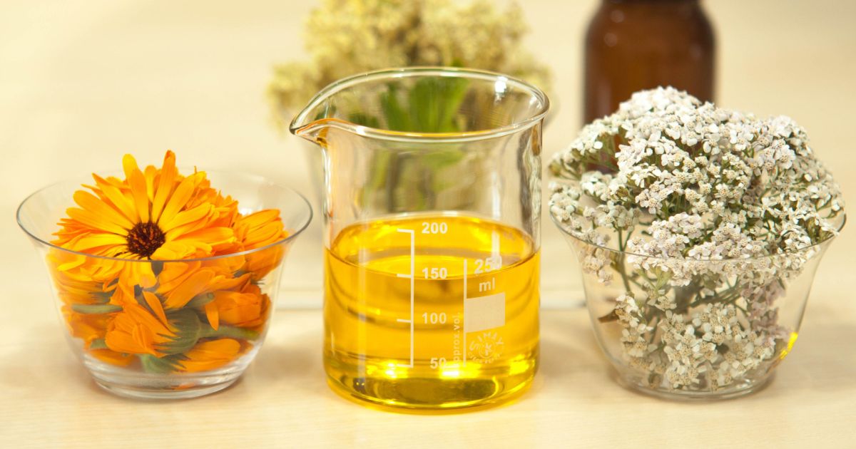 A clear glass beaker filled with a yellow liquid, marked with measurement lines up to 250 ml, is placed in the center. To the left of the beaker is a glass bowl containing bright orange flowers. To the right of the beaker is another glass bowl filled with small white flowers. In the background, there is a brown bottle and some blurred flowers