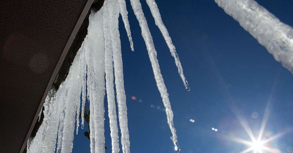 Close-up view of long, sharp icicles hanging from the edge of a roof against a clear blue sky with the sun shining brightly. Winter weather, freezing temperatures, ice formation, cold climate, icicles on roof, winter safety, home maintenance, winter scenery, outdoor winter photography, winter hazards, icicle danger, winter season, cold weather conditions, wintertime beauty.