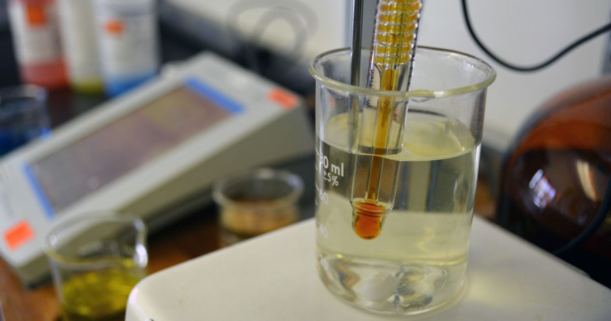 A laboratory setup showing a beaker filled with a clear liquid and a thermometer immersed in it. In the background, there are various laboratory equipment and containers, including a digital device with a screen and other small beakers with different colored liquids.